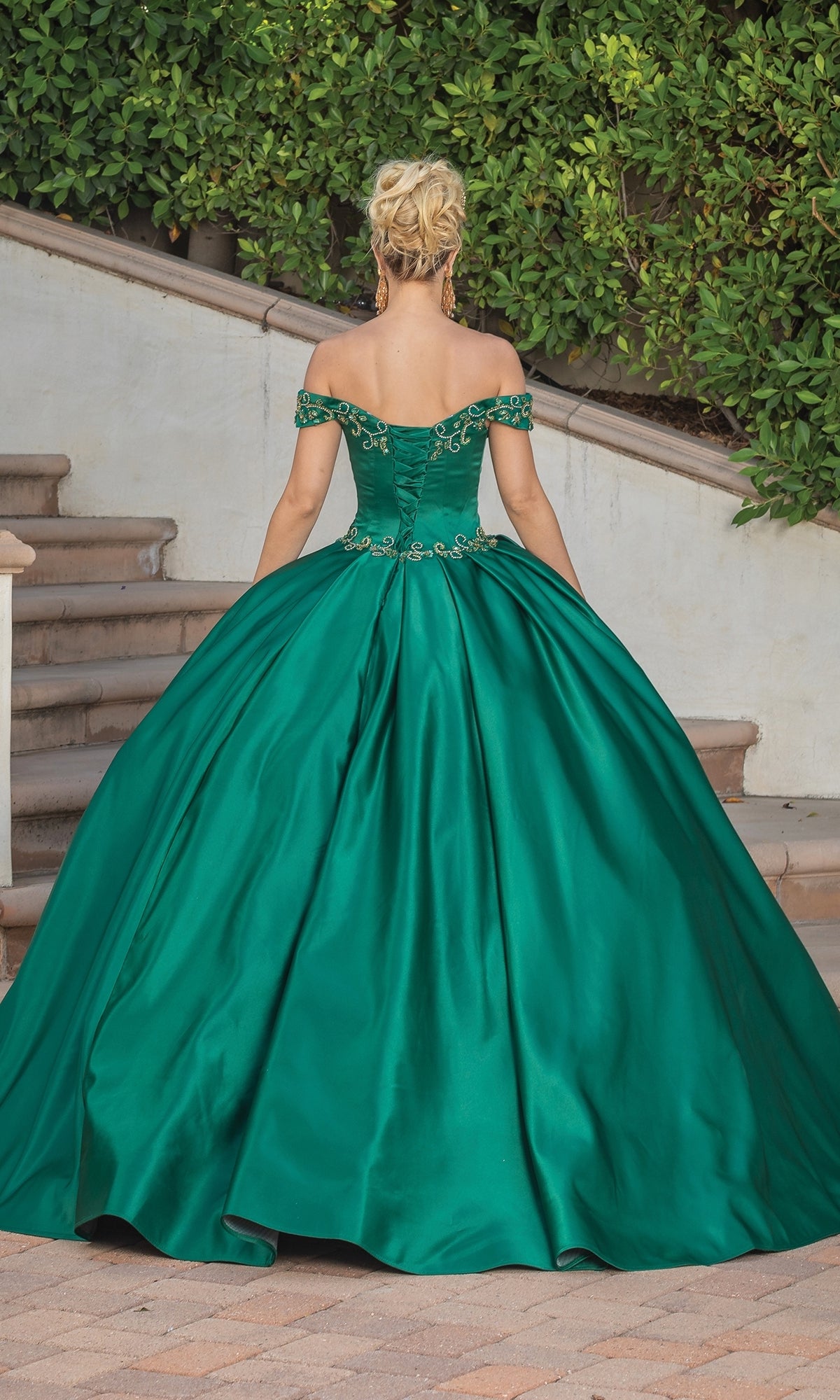 A woman stands outdoors with her back to the camera, wearing the Quinceanera Dress 1466 by Dancing Queen—an emerald green off-the-shoulder a-line ball gown—near steps and lush greenery.